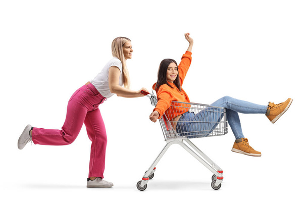 Full length profile shot of a young woman running and pushing a friend inside a shopping cart isolated on white background - Fotoğraf, Görsel
