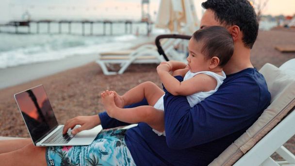 Freelancer man with baby working on laptop near the sea . Remote work concept. work on vacation. 4 k. High quality 4k footage - Fotó, kép