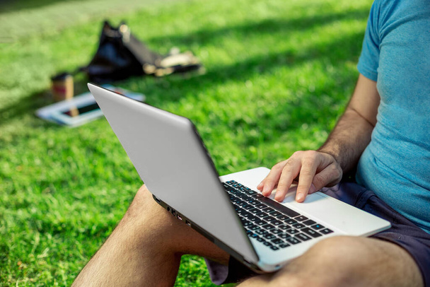 Cropped shot of man using laptop with blank screen while sitting on green grass. Freelance works in the open air in the park - Photo, image