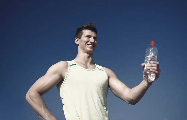 Athlete smiling in green tshirt on blue sky. Man with water bottle in muscular hands. Sport and fitness concept. Bodybuilder with muscles biceps, triceps on sunny day. Healthy lifestyle and health. - Foto, Imagen