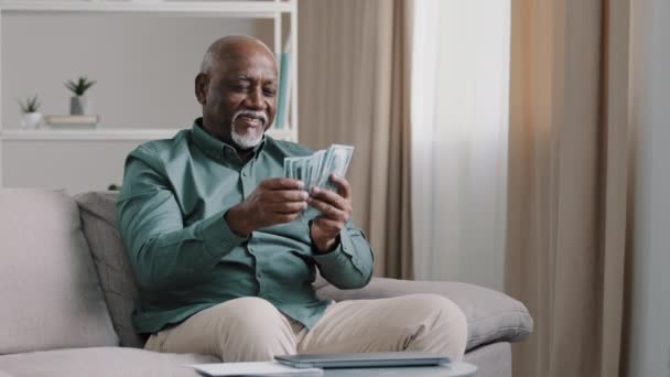 Happy old bald overweight African American senior mature elderly man happy with financial savings salary at home sitting couch. Winning online lottery holding dollars throw up cash money rain falling - Video, Çekim
