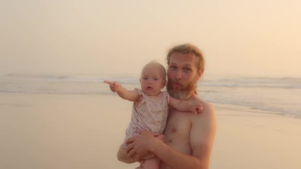 Взрослый кавказский отец с длинными волосами бородатый папочка обнимает маленькую дочку новорожденного ребенка показывая направление пальцем глядя вокруг на небо золотой закат летний день на тропическом пляже - Кадры, видео