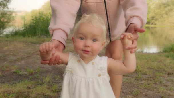 Close-up little Caucasian smiling baby girl infant toddler child kid daughter walking outdoors learning walk in park forest with help of mother kid making first steps grows exploring world develop - Filmagem, Vídeo