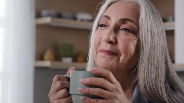 Dreamy inspired old blonde grey-haired woman granny grandmother mature middle aged woman pensioner sitting in kitchen drinking coffee tea hot drink healthy liquid from cup daydreaming relaxing at home - Footage, Video