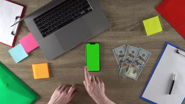 Man is scrolling the pages on the phone with green screen mockup. Vertical position of the phone. 100 dollar bills on the table. - Metraje, vídeo