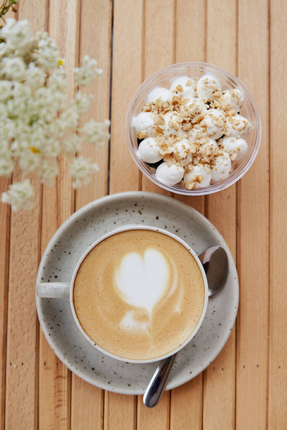 Ceramic white cup of cappuccino with heart, vegan portion of nuts dessert in a disposable cup - appetizer, light breakfast on cafe terrace with blurred foreground of chamomile flowers. Vertical photo. - Foto, imagen