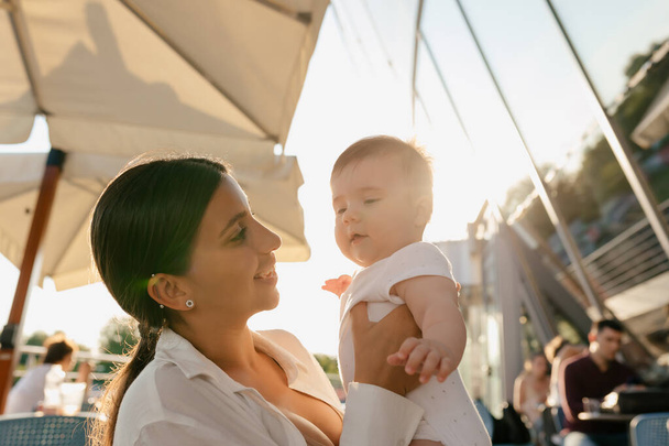 Outdoor photo of young woman with dark hair. tanned skin and lovely smile is holding her little son on hands. Happy smiling european woman playing outdoor in city with her child in sunshine. - Foto, Bild