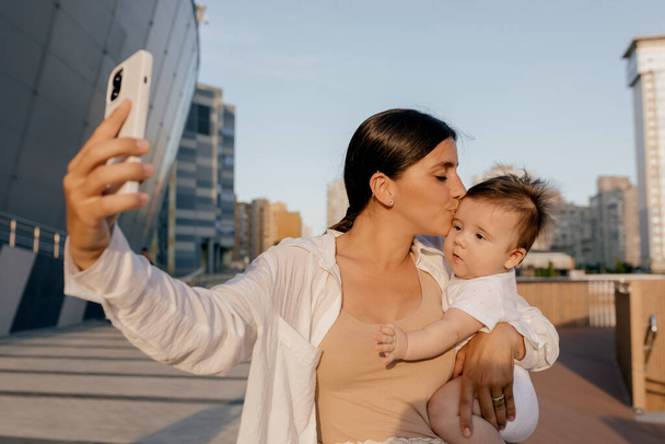 Outdoor photo of brunette woman wiring casual clothes making selfie on smartphone while kissing her little son on city background in sunny warm summer evening  - Foto, imagen
