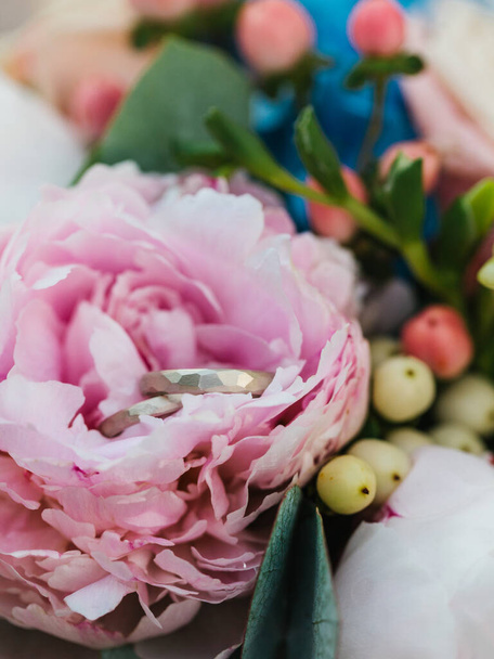 Wedding rings lie in a pink peony bud. In the background are flowers, petals, and berries in a bouquet. - Fotoğraf, Görsel