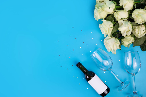 Top view photo of romantic evening, dinner decor hearts love wineglasses with confetti wine bottle and white tape with large bouquet of white roses on isolated blue background with copyspace - Fotó, kép