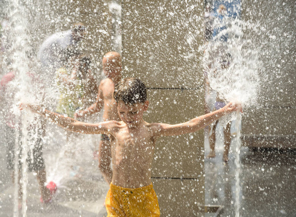 Boy having fun in water fountains. Child playing with a city fountain on hot summer day. Happy kids having fun in fountain. Summer weather. Active leisure, lifestyle and vacation. - Photo, image