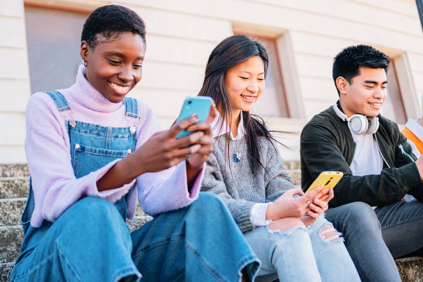 Трое друзей общаются со смартфонами, сидят на улице и улыбаются. высокое качество фото - Фото, изображение