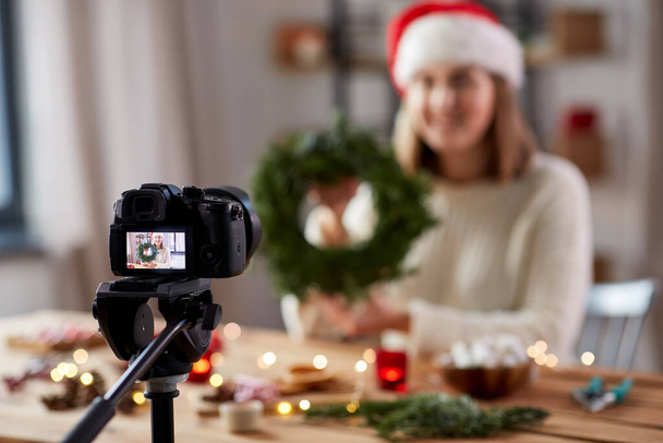winter holidays, diy and video blogging concept - close up of camera recording happy smiling woman or blogger in santa hat making fir christmas wreath at home - Φωτογραφία, εικόνα