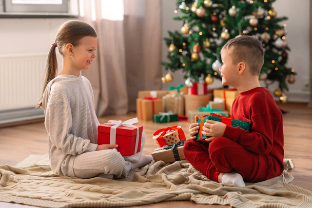 christmas, winter holidays and childhood concept - happy girl and boy in pajamas with gifts sitting on floor in front of each other at home - Photo, image