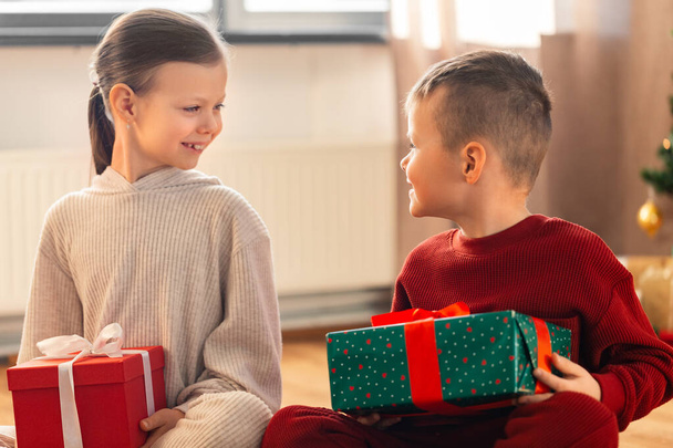 christmas, winter holidays and childhood concept - happy girl and boy in pajamas with gifts sitting on floor in front of each other at home - Zdjęcie, obraz