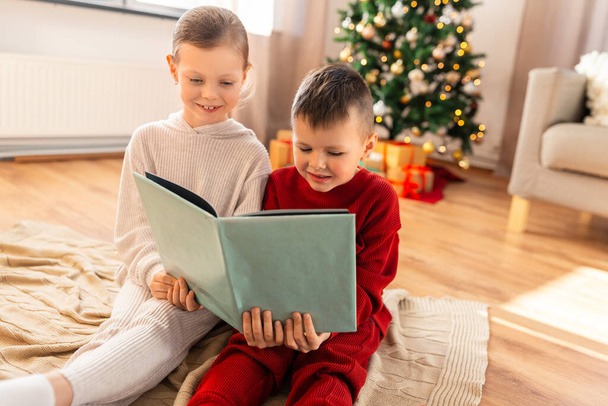 christmas, winter holidays and childhood concept - happy girl and boy in pajamas reading book together sitting on floor in front of each other at home - Φωτογραφία, εικόνα
