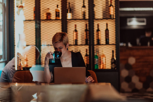 Businesswoman sitting in the coffee shop at wooden table, drinking coffee and using a laptop. Businesswoman browsing internet, chatting, blogging. Selective focus. High quality photo - 写真・画像