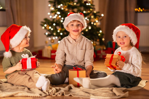 christmas, winter holidays and childhood concept - happy little girl and boys with gifts sitting on floor at home - 写真・画像