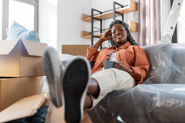 moving, people and real estate concept - happy smiling woman with boxes sitting on sofa with cup of coffee at new home - 写真・画像