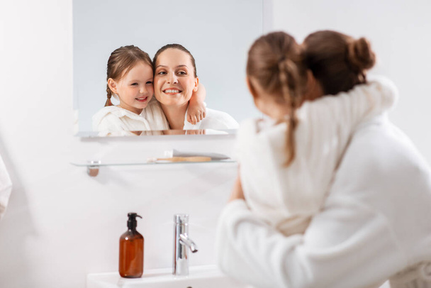 beauty, family and people concept - happy smiling mother and little daughter looking to mirror and hugging in bathroom - Fotó, kép