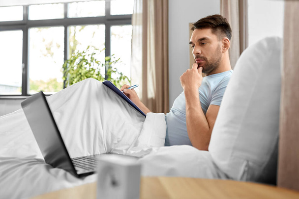 люди, технологии и концепция удаленной работы - человек с ноутбуком и буфером обмена, работающий в постели в домашней спальне - Фото, изображение