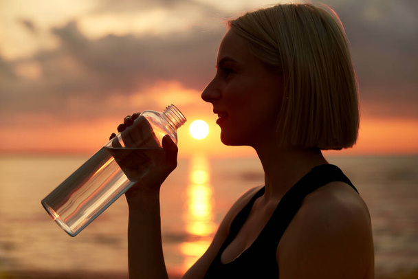 fitness, sport, and healthy lifestyle concept - close up of woman drinking water from bottle on beach over sunset - Foto, Imagem