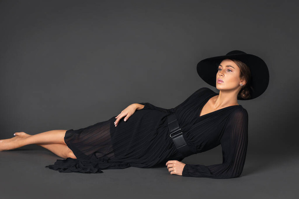 Portrait of an elegant young white woman with wavy hair and beautiful makeup laying by herself on the floor inside a studio with a grey background wearing a black dress with a belt and hat. - 写真・画像