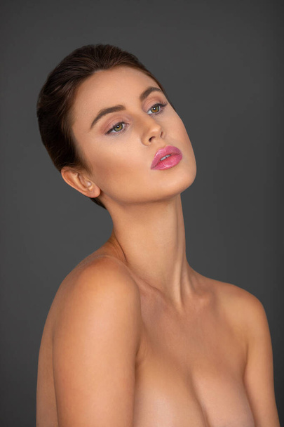 Portrait of a serene young white woman with wavy hair in a bun and beautiful makeup and lipstick posing by herself inside a studio with a grey background. - Foto, afbeelding