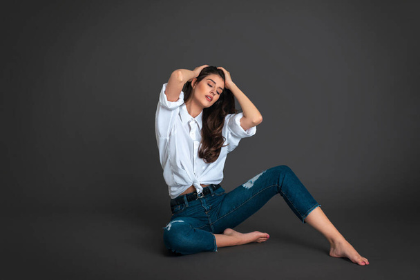 Portrait of a serene latin woman with long dark hair and beautiful makeup sitting by herself on the floor inside a studio with a grey background wearing blue jeans with a white button-up shirt. - Photo, image