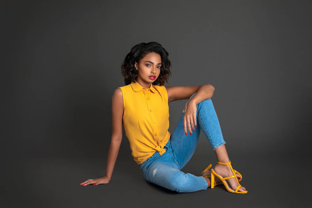 Portrait of a young black woman with curly dark hair and beautiful makeup sitting by herself on the floor inside a studio with a grey background wearing blue jeans with a yellow button-up shirt. - Zdjęcie, obraz