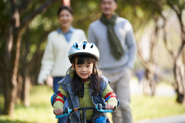 pequeño asiático chica con casco y completo protección engranajes a caballo bicicleta en ciudad parque con padres viendo desde detrás - Foto, imagen