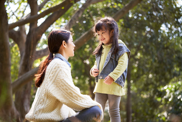 νεαρή ασιάτισσα μητέρα κάθεται στο γρασίδι στο πάρκο έχοντας μια ευχάριστη συνομιλία με χαριτωμένο κόρη - Φωτογραφία, εικόνα