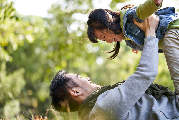 Asyalı baba çimlere uzanıp kızı şehir parkında havaya kaldırırken iyi vakit geçiriyor. - Fotoğraf, Görsel