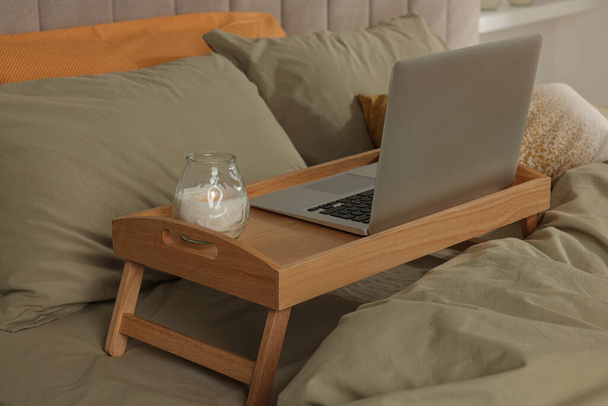 Wooden tray with modern laptop and burning candle on bed indoors - Фото, зображення