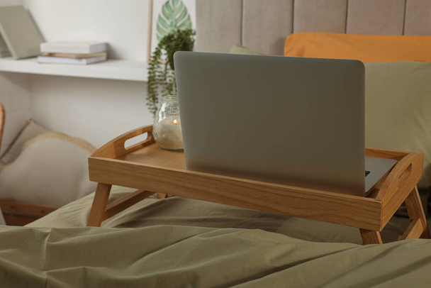 Wooden tray with modern laptop and burning candle on bed indoors - Fotografie, Obrázek