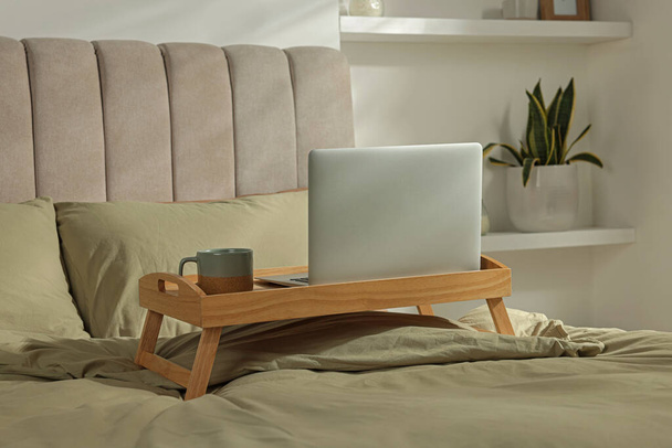 Wooden tray with modern laptop and cup of aromatic drink on bed indoors - Foto, Bild