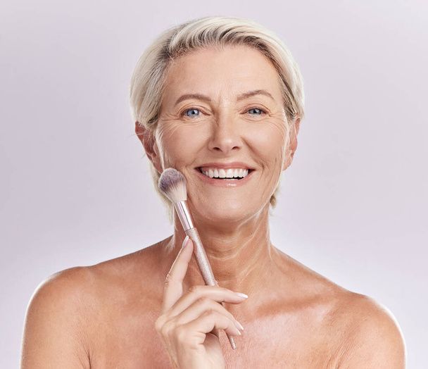 Beauty, skincare and makeup with a senior woman applying cosmetics to her face in studio against a purple background. Head portrait of a female with blusher looking beautiful, happy and healthy. - Foto, Imagem