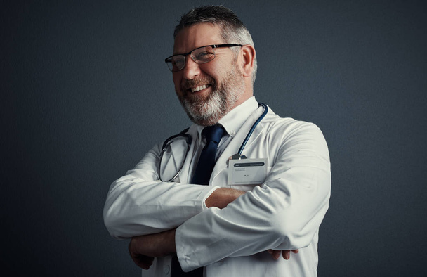 Hes positive about your prognosis. Studio shot of a handsome mature male doctor looking thoughtful while standing with his arms folded against a dark background - Valokuva, kuva