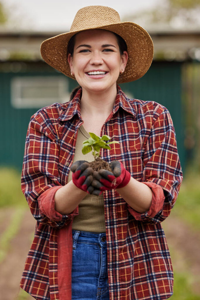 Устойчивый фермер держит в руках растение или саженцы на открытом воздухе, улыбаясь и радуясь своей органической ферме или саду. Молодая женщина-активистка, которая страстно любит устойчивое развитие, стоящая на сельскохозяйственных угодьях. - Фото, изображение