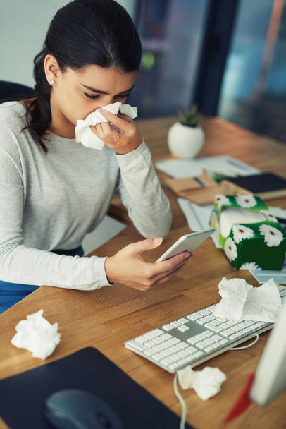 How will I get through this day feeling this terrible. a young businesswoman blowing her nose while texting on a cellphone in an office - Foto, Bild