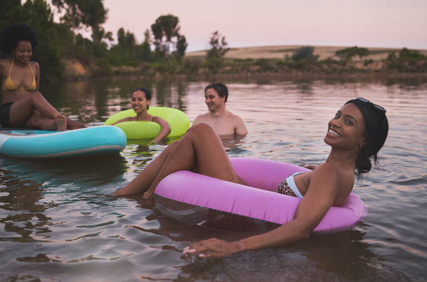 Διακοπές, κολύμπι και φιλία διασκεδάζοντας σε μια λίμνη απολαμβάνοντας το καλοκαιρινό ηλιοβασίλεμα. Χαρούμενοι φίλοι χαλαρώνουν και επιπλέουν στο νερό ενώ μιλούν και δένονται σε διακοπές διαφυγής. - Φωτογραφία, εικόνα