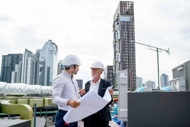 Two engineers or technician man discuss together using building plan or drawing and they also stay on rooftop area of construction site with high building in city as background. - Fotoğraf, Görsel
