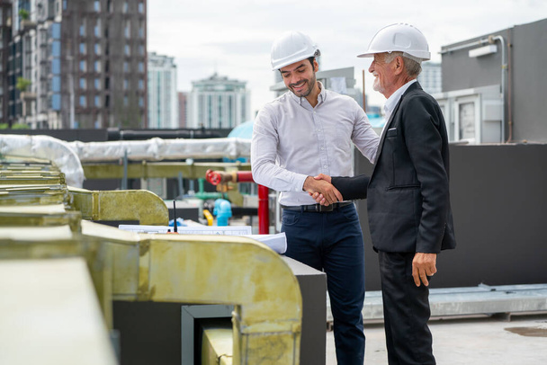 Young engineer shake hands with senior coworker and stay in front of line of air vent in construction site to express success of the project. - Photo, image