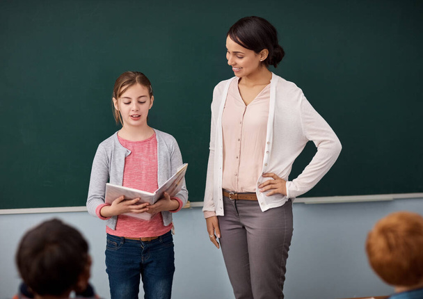 Είναι σίγουρη ομιλήτρια. ένα κορίτσι του δημοτικού διαβάζοντας μπροστά από την τάξη, ενώ στέκεται δίπλα στο δάσκαλό της - Φωτογραφία, εικόνα