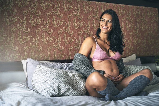 С надеждой смотреть в будущее. Полный снимок привлекательной молодой беременной женщины, сидящей дома на кровати - Фото, изображение