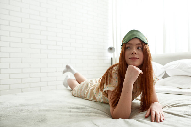 Young focused caucasian girl lying and looking away on bed at home in morning time. Beautiful red haired teen female zoomer wearing homewear and sleep mask. Domestic lifestyle. Modern spacious bedroom - Fotoğraf, Görsel