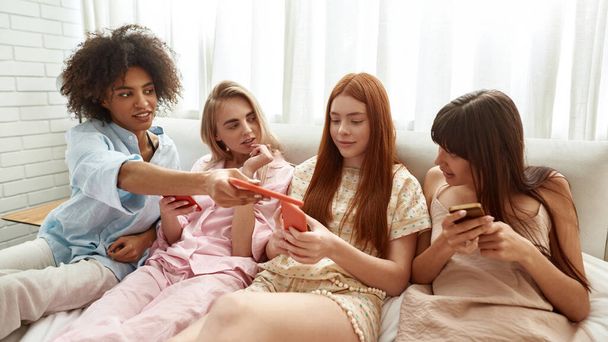 Black girl giving smartphone to girlfriend while they using mobile phones with girlfriends on bed during girlish pajama party at home. Young multiracial female zoomers. Friendship. Rest and leisure - Photo, Image
