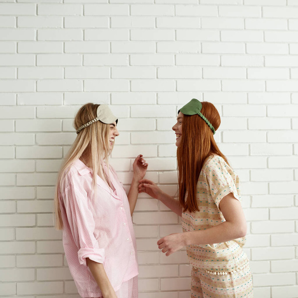 Side view of young caucasian laughing girls looking at each other on wall background at home. Teen female zoomers wearing homewear and sleep masks resting and spending time together. Friendship - Foto, afbeelding