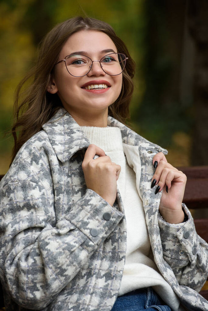 European elegant young woman in a stylish trench coat in a white sweater and blue jeans resting on the bench. warm autumn day. brown wooden bench - Fotoğraf, Görsel