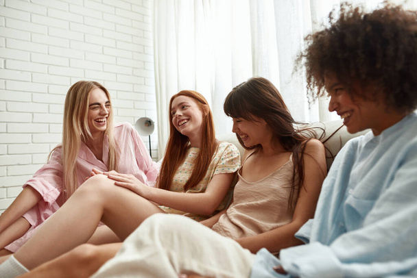 Young multiethnic girlfriends laughing and enjoying time on bed during girlish pajama party at home. Black and caucasian zoomer girls resting together. Friendship. Bedroom in modern apartment - Fotó, kép